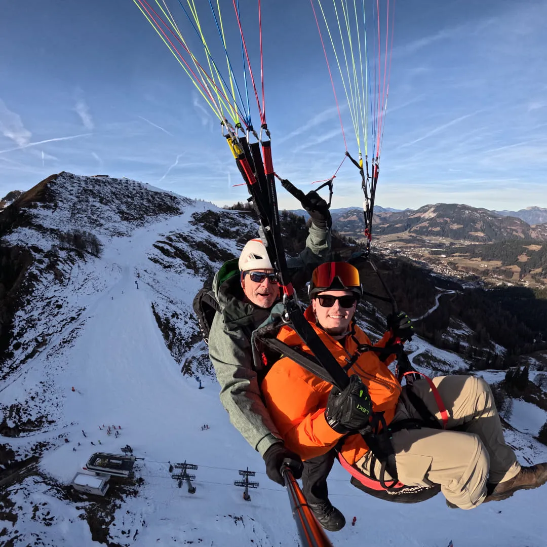 Zwei Personen beim Gleitschirmfliegen über schneebedeckter Landschaft.