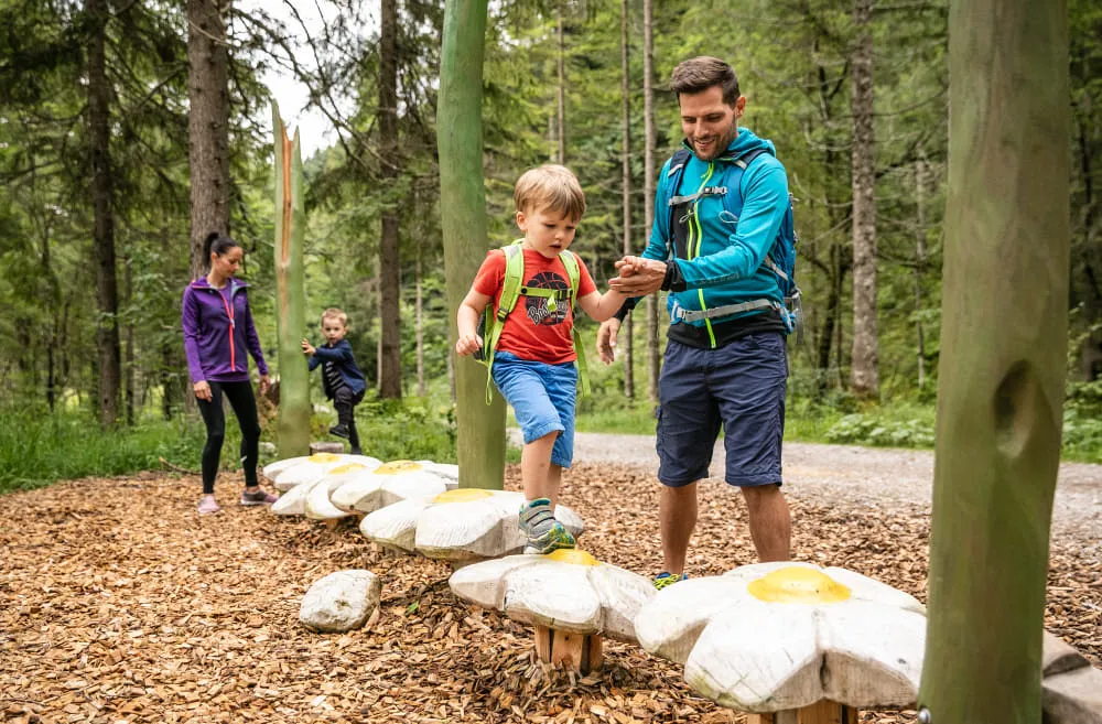 Familie wandert im Wald, Kind springt über Holzsteine.